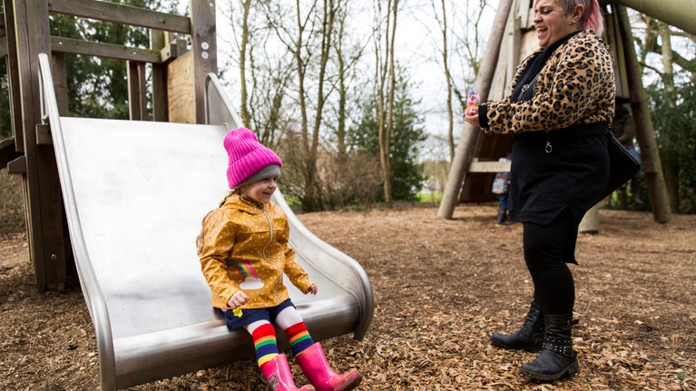 A child wearing winter coat, hat and wellies goes down a wide metal slide, as a parent cheers her on